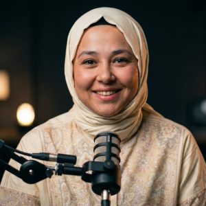 Krem rengi başörtüsüyle gülümseyen Semanur, loş ışıklı podcast stüdyosunda mikrofonun önünde.