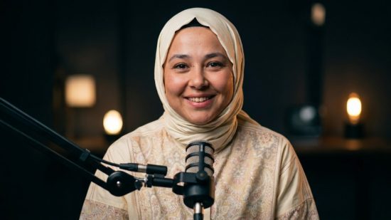 Krem rengi başörtüsüyle gülümseyen Semanur, loş ışıklı podcast stüdyosunda mikrofonun önünde.